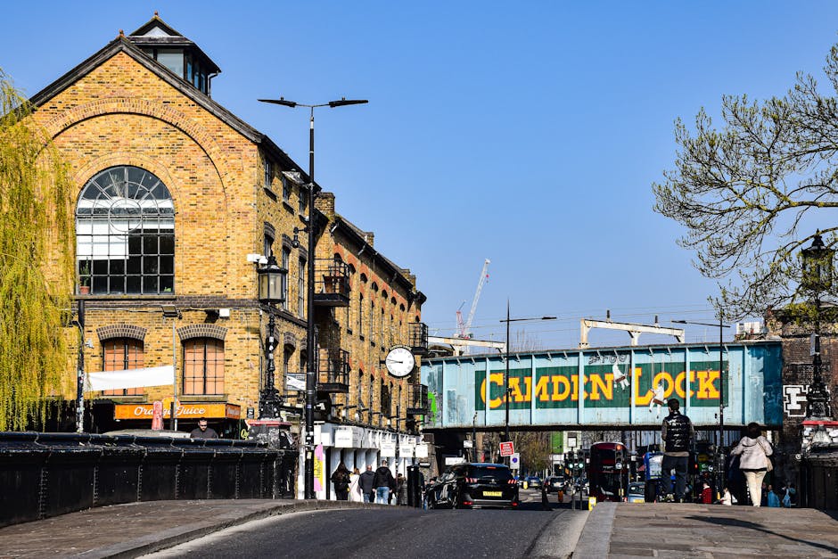 A lively urban street scene in Lea Bridge featuring a mix of historic brick buildings with large arched windows and modern structures. In the foreground, the street is lined with pedestrians walking a
