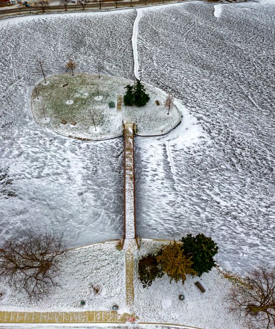 An aerial view of a snow-covered communal courtyard showing a pathway leading from the bottom center to a small, central landscaped area with a few small trees and bushes, surrounded by covered seatin